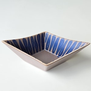 May include: A square ceramic bowl with a light brown exterior and a blue and white patterned interior. The bowl has a geometric design with a slightly raised rim. The interior features vertical blue stripes with white accents.