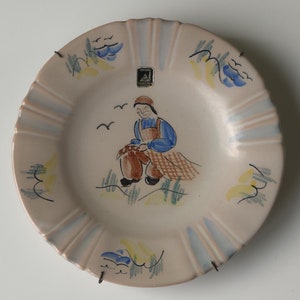 Puede incluir: Plato de cerámica decorativo con borde festoneado. El plato presenta una escena pintada a mano de una persona pescando, con detalles en azul, amarillo y marrón. El plato está montado con colgadores de alambre metálico.