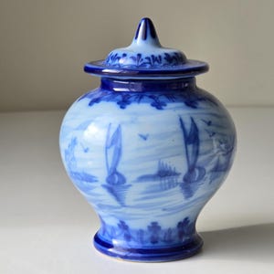 Op de afbeelding: Een kleine, blauw-witte keramische pot met deksel. De pot heeft een handgeschilderde landschapsscène met zeilboten. Het deksel en de basis zijn donkerder blauw en de pot is ongeveer 13 cm hoog.
