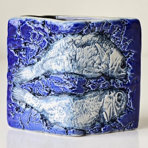 Puede incluir: Un jarrón cuadrado de cerámica con exterior azul texturizado. Dos peces blancos en relieve en la parte delantera. El jarrón es rectangular y probablemente decorativo. Medidas en centímetros.