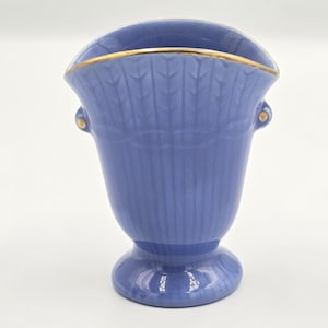 Puede incluir: Un jarrón azul bígaro con borde dorado y pequeños detalles dorados. El jarrón tiene un diseño acanalado y una base de pedestal. El jarrón es probablemente de cerámica o porcelana y es adecuado para exhibir flores o como objeto decorativo.