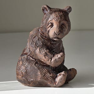 Può includere: Statuetta di orso in ceramica marrone in posizione seduta. L'orso ha una texture di pelliccia dettagliata ed è seduto con le zampe giunte. L'oggetto è decorativo.