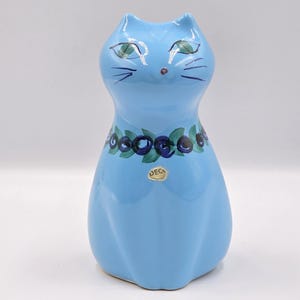 Puede incluir: Figura de gato de cerámica azul claro con ojos verdes y cara pintada en blanco y negro. El gato tiene un collar decorativo de bayas azules y hojas verdes. Hay una pequeña pegatina redonda dorada.