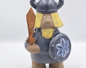 May include: A ceramic Viking figurine with a blue helmet and tunic, holding a wooden sword and a shield with a floral design. The figurine has yellow hair and brown boots. The overall design is whimsical.