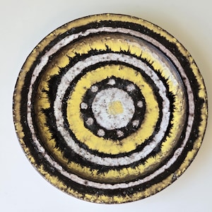 Puede incluir: Un plato redondo de cerámica con un diseño de círculos concéntricos en negro, blanco y amarillo. El centro del plato tiene un círculo blanco con puntos negros alrededor.