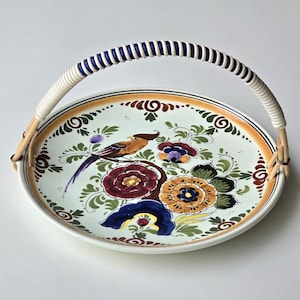 Peut inclure: Un plateau de service rond en céramique peint à la main avec une poignée tressée. Le plateau présente un motif d'oiseau et de fleurs dans des tons d'orange, de bleu, de violet et de vert. La poignée est blanche avec des rayures bleues.