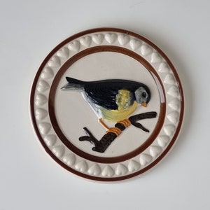 Puede incluir: Un plato de cerámica blanco con un borde marrón y un patrón de corazones blancos. El plato presenta una imagen pintada de un pájaro amarillo y negro posado en una rama marrón.