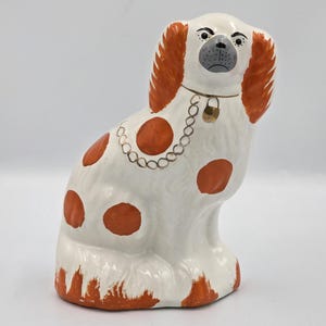 May include: A white ceramic dog figurine with orange spots, long orange ears, and a painted grey muzzle. The dog features a painted gold chain necklace with a small gold padlock charm around its neck, and orange accents at the base.