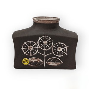 Puede incluir: Un jarrón de cerámica negro con un diseño floral blanco estilizado. El jarrón tiene una base ancha y plana y un cuello estrecho. El jarrón está marcado con una pegatina amarilla que dice "EST".