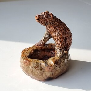 Puede incluir: Un cenicero de cerámica con una figurita de oso marrón en la parte superior. El cenicero es marrón y tiene un esmalte moteado.