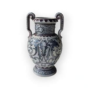 Può includere: Un vaso in ceramica grigio e bianco con un motivo floreale e due manici. Il vaso ha un bordo e una base di colore viola scuro.