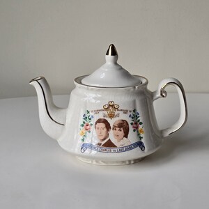 Puede incluir: Tetera de cerámica blanca con tapa y asa doradas. La tetera presenta una imagen conmemorativa de la boda del príncipe Carlos y Lady Diana Spencer con el texto "To Commemorate the Marriage of HRH Prince Charles to Lady Diana Spencer, July 29th 1981."