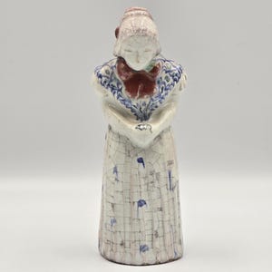 Peut inclure: Figurine en céramique représentant une femme en costume traditionnel hollandais. La figurine porte une robe blanche avec des touches de bleu, un nœud rouge et un châle à motifs bleus et blancs. Le visage de la figurine est baissé.