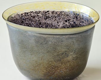 May include: A handcrafted, metallic grey bowl with a textured surface. The rim is a translucent yellow, and the interior appears to be filled with a dark, speckled material. The bowl has a rounded shape and sits on a plain background.