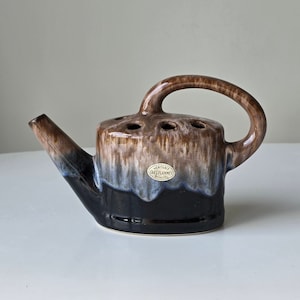 Könnte beinhalten: Eine braune und schwarze Keramik-Gießkanne mit blauer Glasur. Die Gießkanne hat einen Griff und einen Ausguss. Sie trägt ein Etikett mit der Aufschrift "VERITABLE GRÈS FLAMMÉ GRANITO".