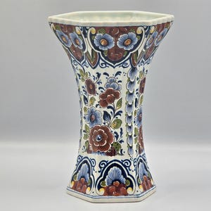 May include: A tall, hexagonal vase with a flared top. The ceramic vase features a white background with a repeating floral pattern in blue, red, and green. The rim is a dark red with blue floral accents.