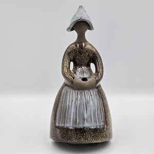 Puede incluir: Figura de cerámica de una mujer con traje tradicional holandés. La figura tiene un vestido marrón y dorado moteado, un delantal blanco y un sombrero azul claro. La figura está de pie con las manos juntas.