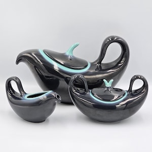 May include: A set of three black ceramic serving dishes. The set includes a gravy boat, a covered sugar bowl, and a serving dish. Each piece has a turquoise accent and handle.