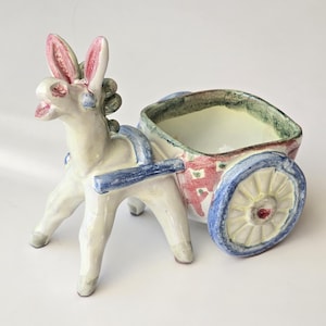 Könnte beinhalten: Eine skurrile Keramik-Esel-Figur, die einen Wagen-Pflanztopf zieht. Der Esel ist weiß mit rosa gefütterten Ohren und blauen Geschirrdetails. Der Wagen hat rote und blaue Blumenmuster und ein blaues Rad.
