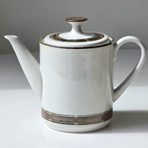 Könnte beinhalten: Eine weiße Teekanne aus Keramik mit einem braunen Deckel und einem braunen geometrischen Muster um die Basis.