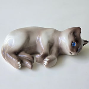 Op de afbeelding: Een keramieken beeldje van een Siamese kat in een liggende houding. De kat heeft een lichtbruin lichaam met donkerbruine aftekeningen op het gezicht, oren en poten. Het heeft helderblauwe ogen en rust op zijn voorpoten.