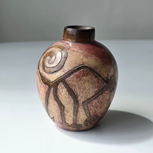 Puede incluir: Un pequeño jarrón redondo de cerámica con un esmalte marrón, rojo y beige. El jarrón está decorado con un diseño estilizado de figuras y una espiral.