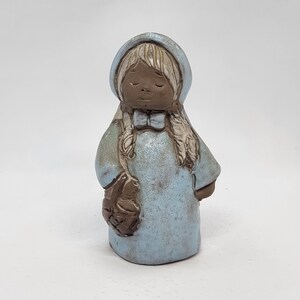 May include: A small ceramic figurine of a girl wearing a blue hooded dress and holding a basket. The figurine is made of brown clay and has a rustic, hand-crafted look.