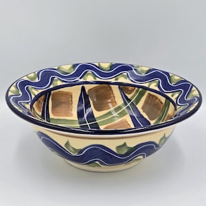 Könnte beinhalten: Eine Keramikschale mit cremefarbenem Äußeren und einem geometrischen Design in Blau, Grün und Braun. Der Rand weist ein wellenförmiges blaues und grünes Muster auf. Das Innere der Schale hat ein gitterartiges Muster mit braunen und grünen Quadraten.
