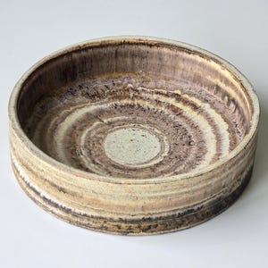 Könnte beinhalten: Eine runde Keramikschale mit breitem, flachem Rand. Die Schale zeigt konzentrische Kreise in verschiedenen Braun-, Beige- und Cremetönen, wodurch ein geschichteter Effekt entsteht. Das Innere hat eine hellere, cremefarbene Farbe. Die Schale hat eine rustikale, erdige Ästhetik.