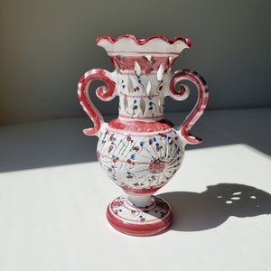 May include: A white ceramic vase with red and pink floral designs and cutouts. The vase has two handles and a scalloped rim.