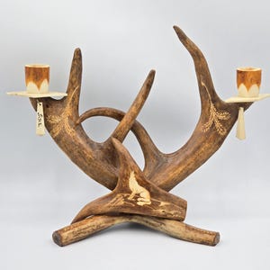 Peut inclure: Un candélabre en bois de cerf marron doté de deux bougeoirs avec des bases carrées plates. Les bois sont ornés de motifs gravés représentant un loup hurlant et du feuillage. Une petite étiquette rectangulaire avec le texte "GRE" est suspendue.