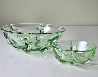 May include: Two light green glass bowls. The larger bowl has three sections and a wavy design. The smaller bowl also has three sections and a similar wavy design. Both bowls are transparent and sit on a white surface.