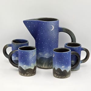 Peut inclure: Un pichet en céramique et quatre tasses avec un motif de ciel nocturne. Les pièces bleues présentent un ciel étoilé, un croissant de lune et un paysage de collines. Les tasses ont des anses et des bords noirs.