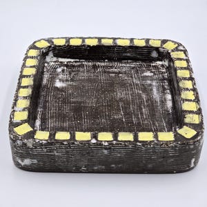 May include: A square, ceramic tray with a dark brown interior and a textured surface. The rim is lined with yellow rectangular accents. The tray has a rustic, handmade appearance.
