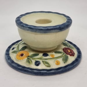Puede incluir: Un portavelas de cerámica blanca con un borde azul y un diseño floral. El portavelas tiene una base redonda y una parte superior en forma de taza con un agujero en el centro para una vela.