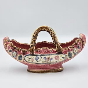Puede incluir: Una cesta de cerámica vintage con asa, con base y borde granate con detalles dorados. La cesta tiene un diseño floral en el centro y elementos decorativos alrededor del borde. El asa es marrón y dorada.