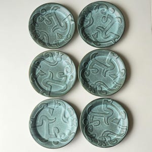 Könnte beinhalten: Sechs teal-farbene Keramikplatten mit erhabenem, abstraktem Design. Die Teller sind rund und haben eine leicht strukturierte Oberfläche. Das Design zeigt fließende Linien und geometrische Formen. Die Teller sind in einem 2x3 Raster angeordnet.