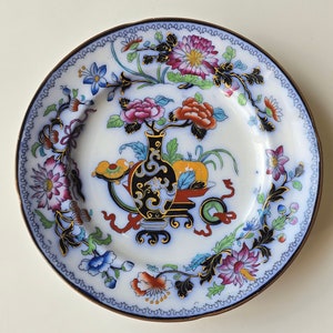 Könnte beinhalten: Ein antiker Porzellanteller mit einem blauen und weißen Blumendesign. Der Teller zeigt ein zentrales Motiv einer Vase mit Blumen und Früchten, umgeben von einem Rand aus stilisierten Blumenmustern.