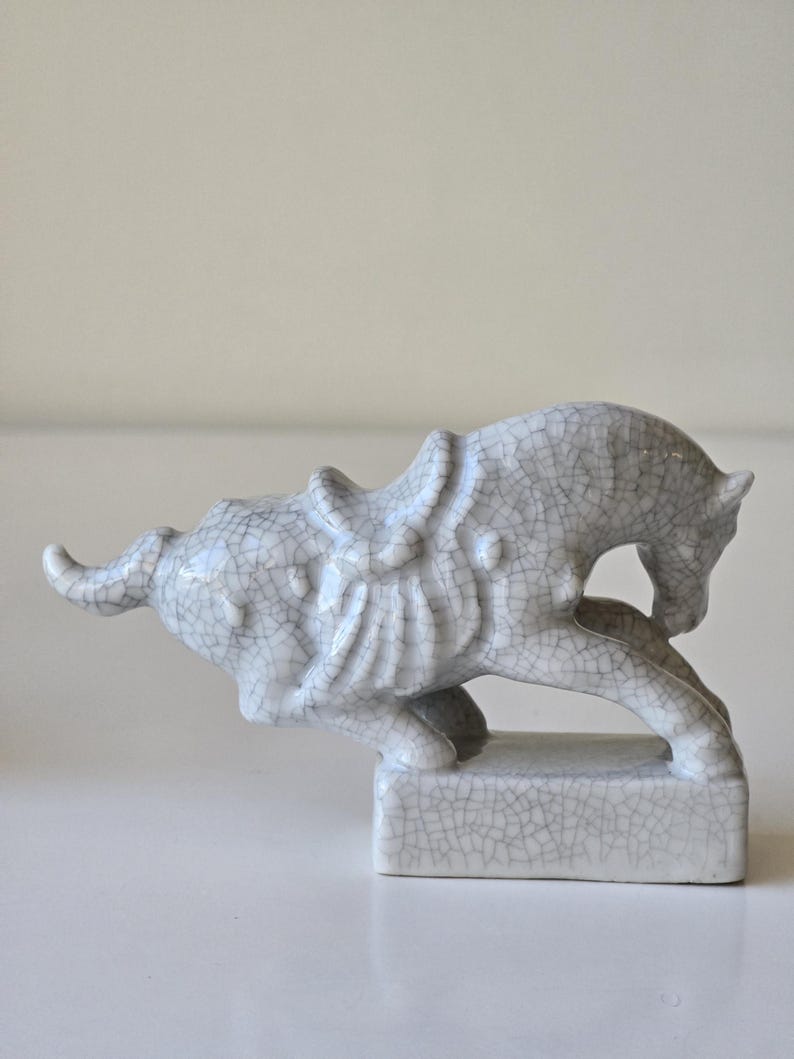 May include: A white ceramic horse figurine with a cracked glaze finish. The horse is in a running pose with its head turned to the side. The figurine is on a square base.