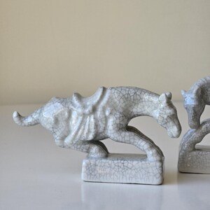 May include: Two white ceramic horse figurines with a crackled glaze finish. The horses are in a running pose and have saddles. The figurines are on a white surface.