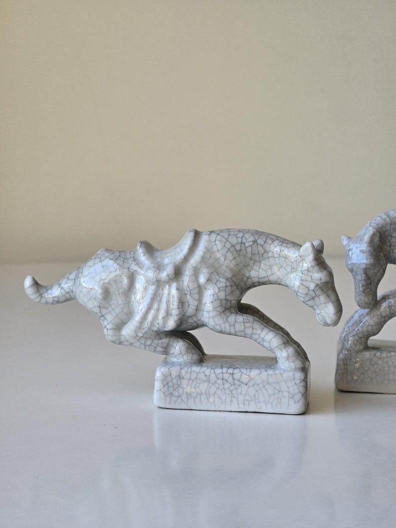 May include: Two white ceramic horse figurines with a crackled glaze finish. The horses are in a running pose and have saddles. The figurines are on a white surface.