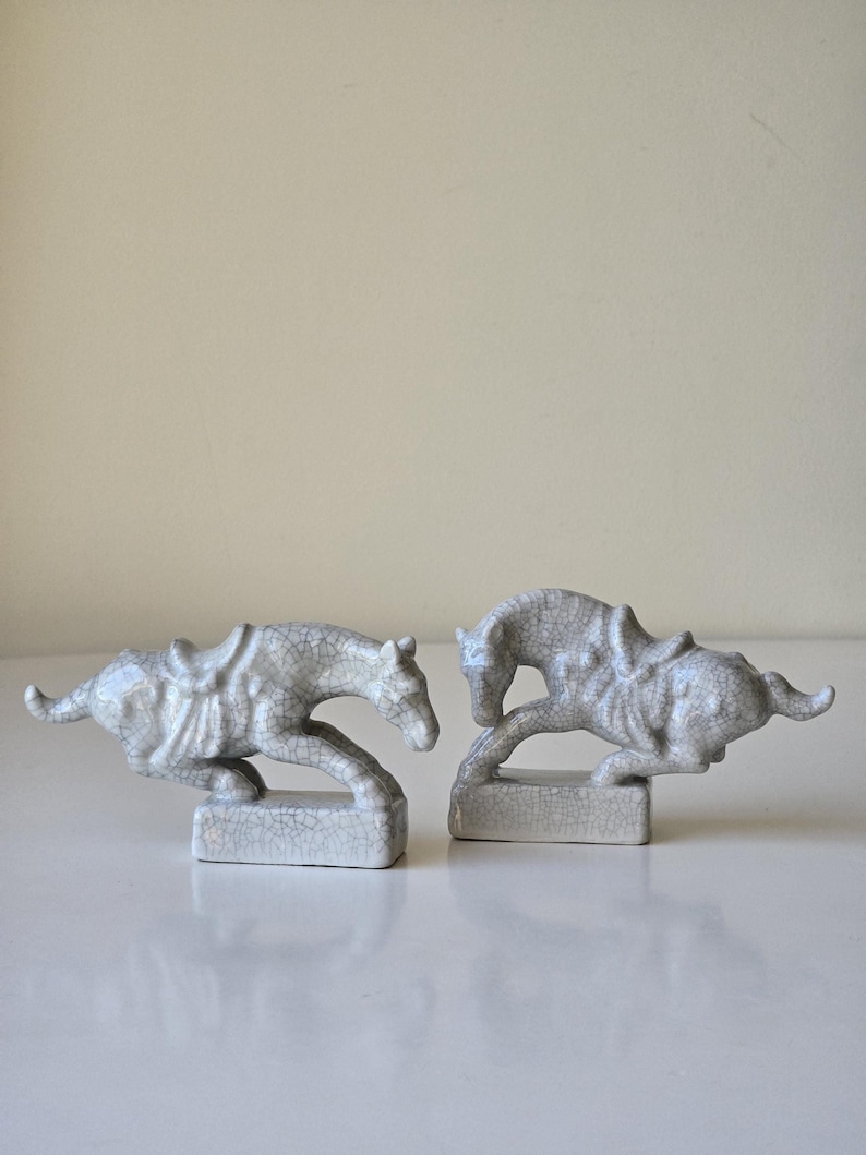 May include: Two white ceramic horse figurines with a crackled glaze finish. The horses are in a running pose and are on small rectangular bases.