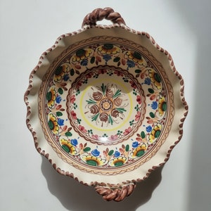 Könnte beinhalten: Eine weiße Keramikschüssel mit einem braunen Seilgriff. Die Schüssel ist mit einem bunten Blumenmuster in den Farben Gelb, Blau, Grün, Rot und Braun verziert.