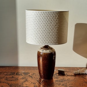 Może przedstawiać: Brązowa ceramiczna lampa stołowa z białym, plecionym abażurem. Podstawa lampy ma ozdobny wzór wokół górnej części.