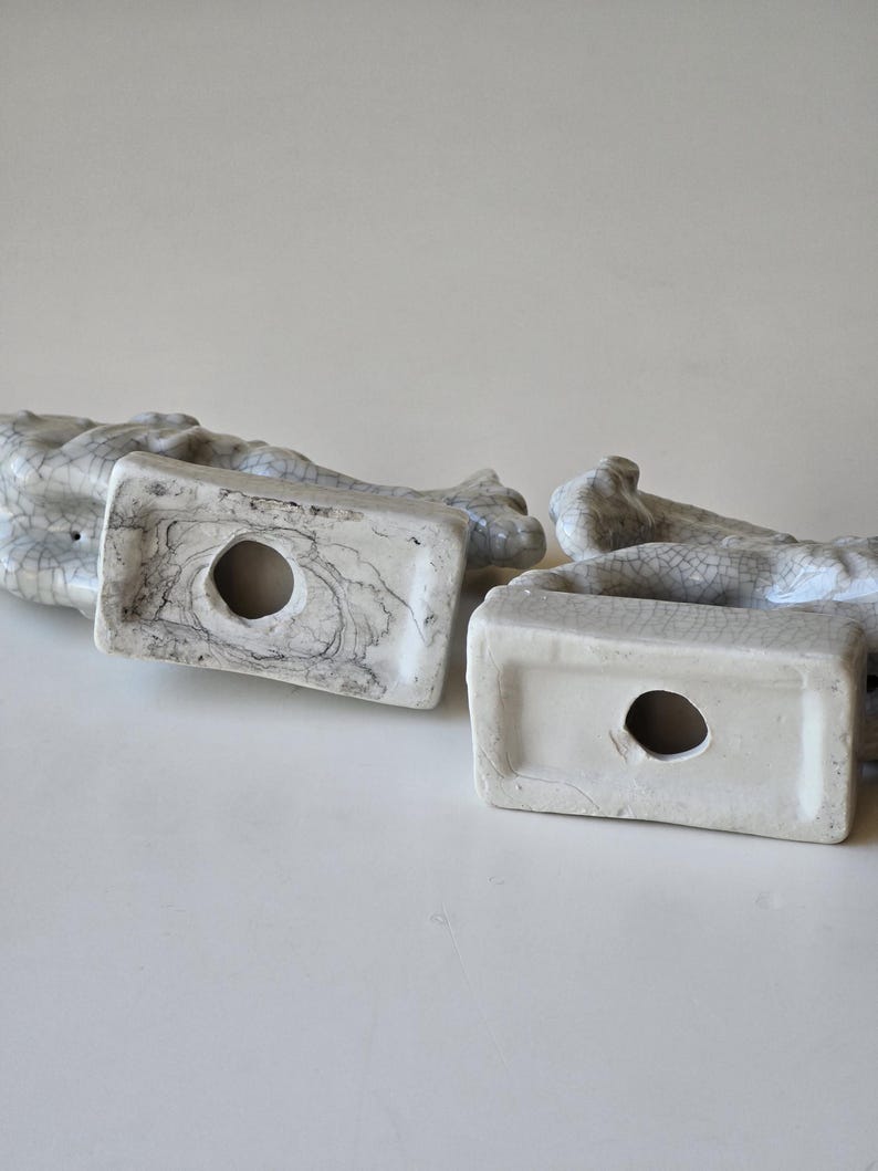 May include: Two white ceramic figurines with a crackled glaze finish. The figurines are in the shape of a seated animal, possibly a dog or cat. The bottom of each figurine has a hole for a candle.