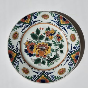 May include: A white ceramic plate with a blue, green and orange floral design. The plate features a central floral motif with yellow, orange and red flowers, surrounded by a border of blue and green swirls and orange and yellow flowers.