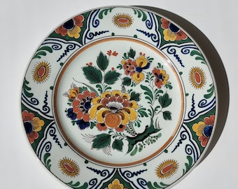Könnte beinhalten: Ein weißer Keramikteller mit einem blauen, grünen und orangefarbenen Blumendesign. Der Teller zeigt ein zentrales Blumenmotiv mit gelben, orangefarbenen und roten Blumen, umgeben von einem Rand aus blauen und grünen Spiralen und orangefarbenen und gelben Blumen.