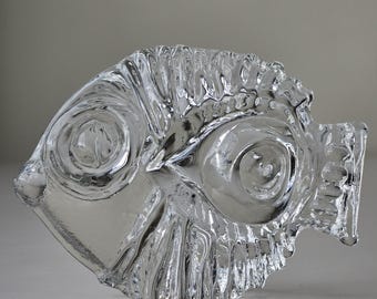 May include: A clear glass sculpture of a fish with a detailed, textured design. The fish has a rounded body and a slightly curved tail.