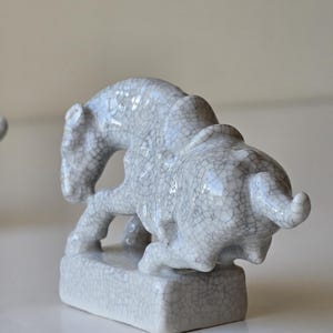 May include: A white ceramic figurine of a mythical creature, possibly a lion or a griffin, with a cracked glaze finish. The figurine is standing on a square base.
