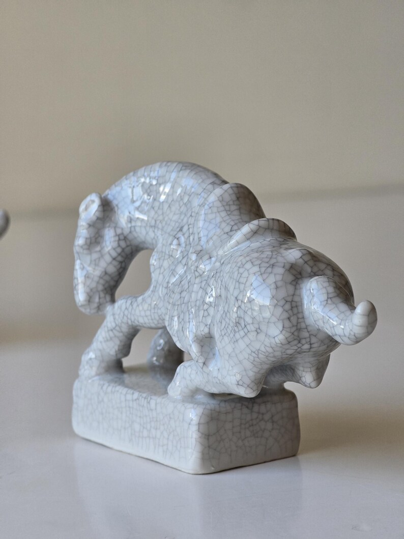 May include: A white ceramic figurine of a mythical creature, possibly a lion or a griffin, with a cracked glaze finish. The figurine is standing on a square base.